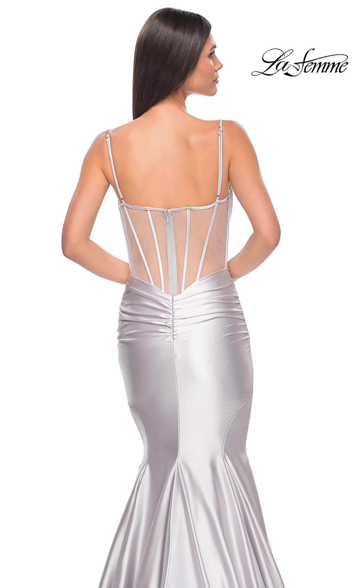 A woman stands with her back to the camera in a form-fitting silver La Femme Long Corset Mermaid Prom Dress 32269, featuring spaghetti straps and a sheer back panel. The